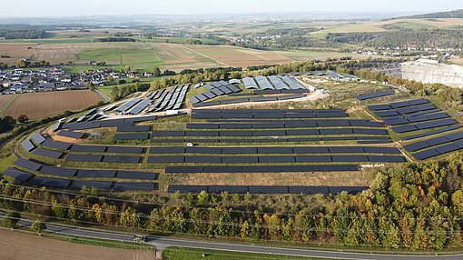 Im Fokus stehen eine große Anzahl an Solarmodulen installiert auf einer größeren Fläche. Im Hintergrund sieht man noch einige Einfamilienhäuser, sowie Ackerflächen